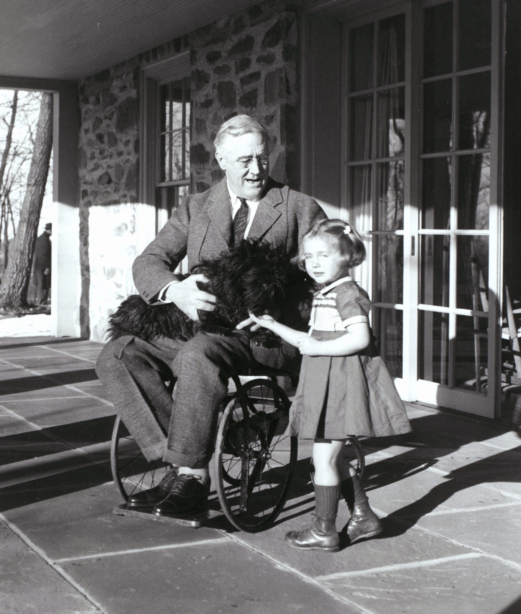 President Roosevelt in his wheelchair on the porch at Top Cottage in Hyde Park, NY with Ruthie Bie and Fala. February 1941.