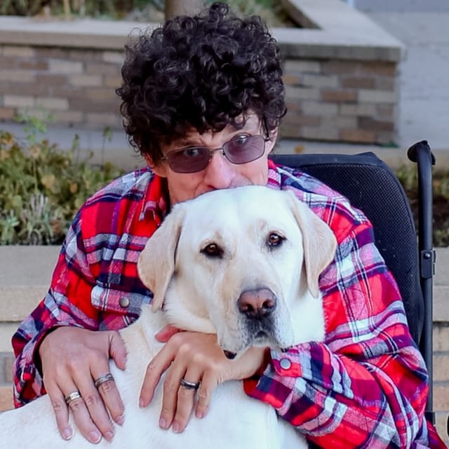A person in a wheelchair wearing a red plaid shirt and sunglasses embraces a large yellow Labrador Retriever, who looks directly at the camera.