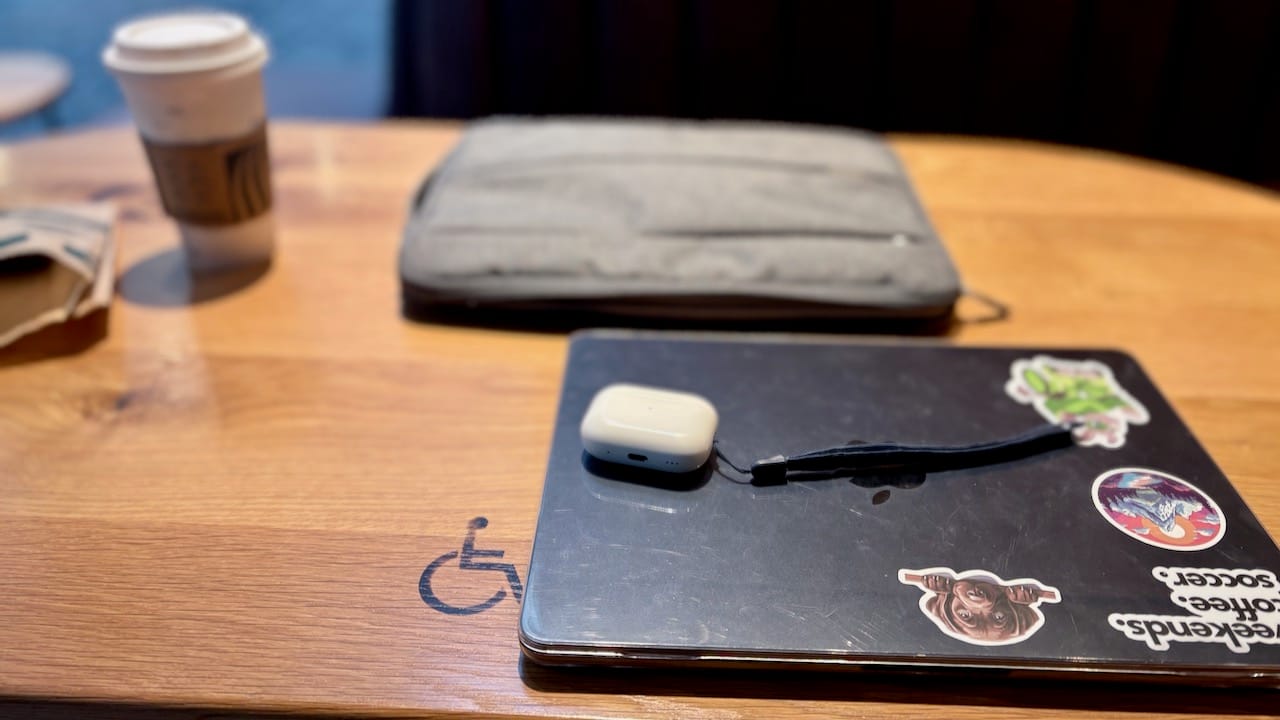 A wooden café table with a laptop covered in stickers, a pair of wireless earbuds, and a coffee cup. The table features a wheelchair accessibility symbol, indicating a designated accessible seating area. A soft-focus background suggests a cozy coffee shop setting.