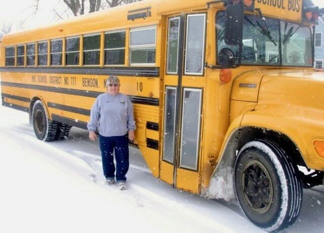Grandma Marlys standing in the snow next to a yellow school bus with ‘Benson School District 777’ written on the side.
