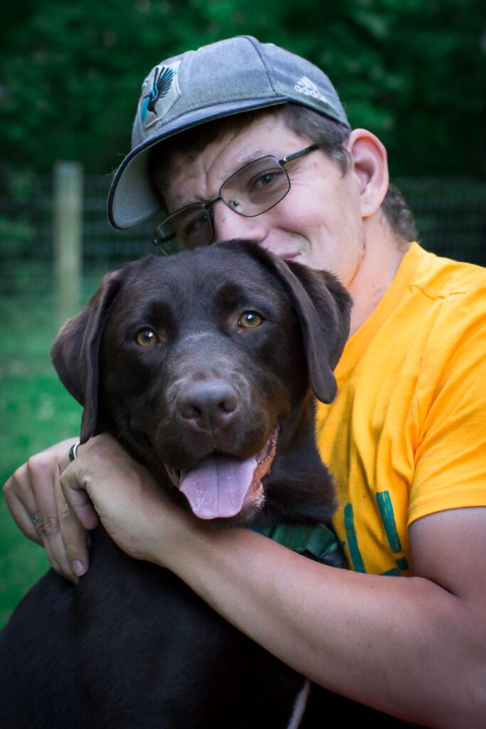 Levi wearing a gray Minnesota United FC hat and yellow shirt, hugging his chocolate Labrador service dog, Dempsey, outdoors with a green background.