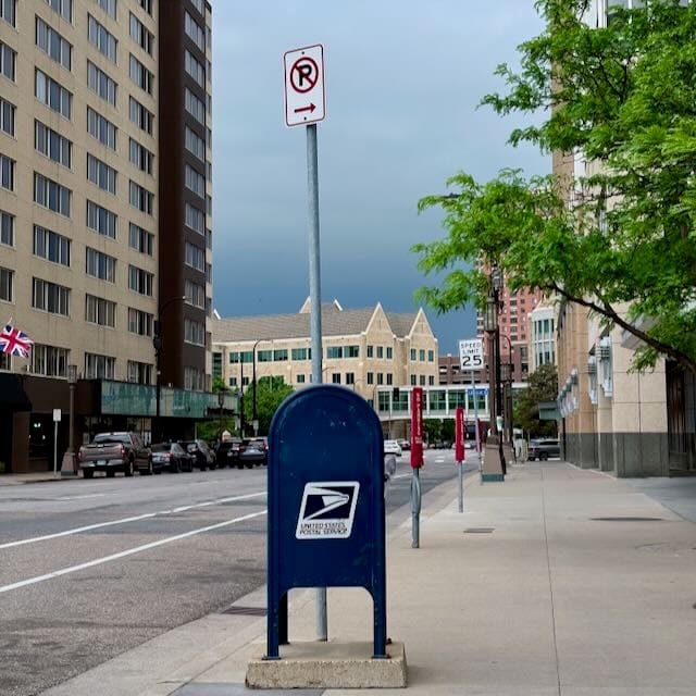 A city street in Minneapolis with dark storm clouds rolling in, a USPS mailbox in the foreground.
