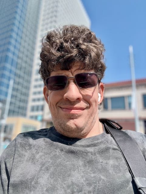 Adult man with curly hair, wearing sunglasses and a dark gray shirt, smiling outside in front of a tall building under a clear blue sky.