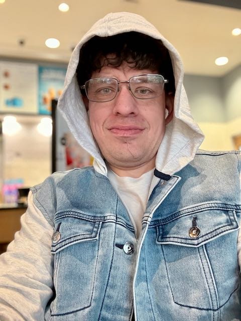Adult man with curly hair wearing clear glasses, a gray hoodie, and a denim vest, sitting indoors and smiling slightly in a well-lit coffee shop.
