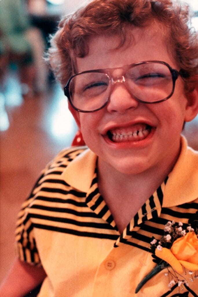 A young boy with curly hair and large glasses smiling brightly at the camera, wearing a yellow and black striped collared shirt with a yellow boutonnière pinned to it.