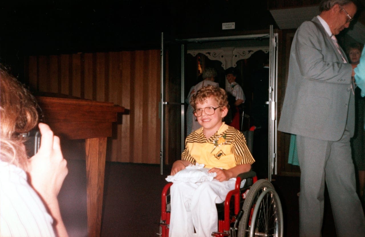 A young boy with curly hair and glasses sits in a red wheelchair, smiling. He’s wearing a yellow and black striped shirt with a boutonnière. An adult and others are visible in the background.