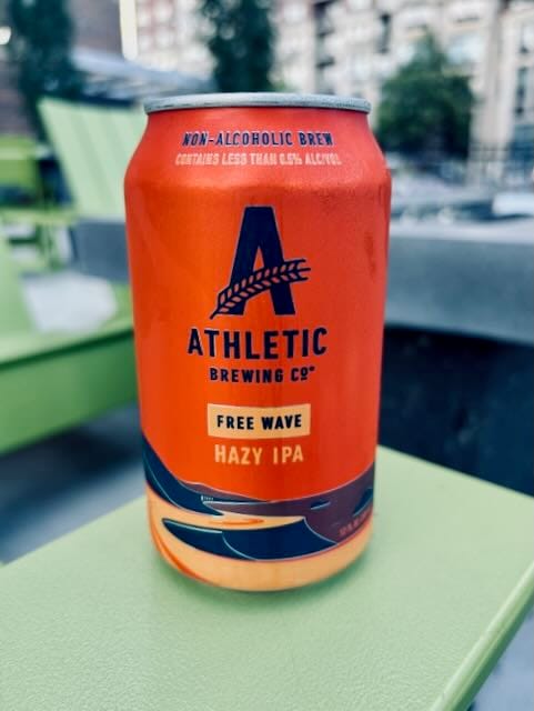 A cold can of Athletic Brewing Co.’s Free Wave Hazy IPA rests on a bright green patio chair, with an urban apartment complex and soft greenery in the blurred background. The vibrant orange can is prominently labeled as a non-alcoholic brew.
